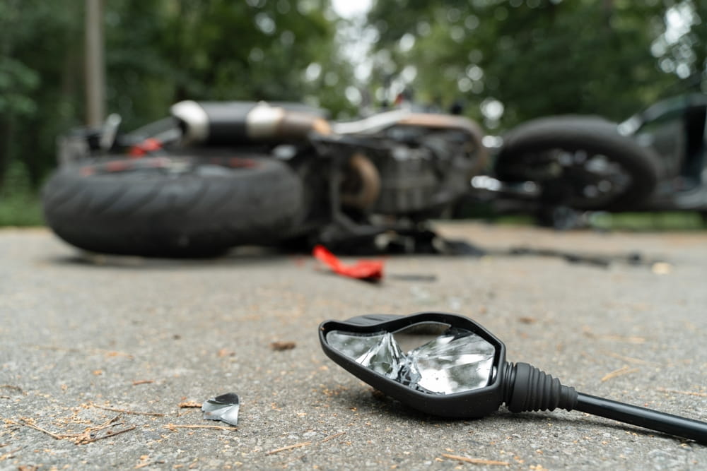 Broken motorcycle mirror in foreground with crashed bike in background illustrating accident damage and insurance settlement gaps in Florida